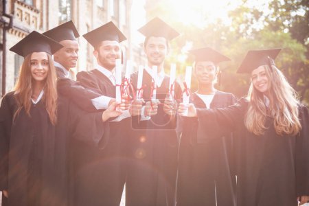 Foto de Los graduados exitosos en vestidos académicos tienen diplomas, miran a la cámara y sonríen mientras están al aire libre. - Imagen libre de derechos