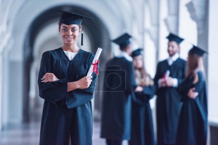 Téléchargez les photos : Diplômés avec succès dans les robes académiques parlent dans la salle universitaire, fille au premier plan est titulaire d'un diplôme, regardant caméra et sourire - en image libre de droit