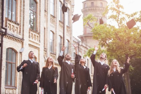 Téléchargez les photos : Les diplômés qui réussissent dans les robes universitaires sont titulaires de diplômes, jetant leurs casquettes et souriant tout en se tenant debout à l'extérieur - en image libre de droit