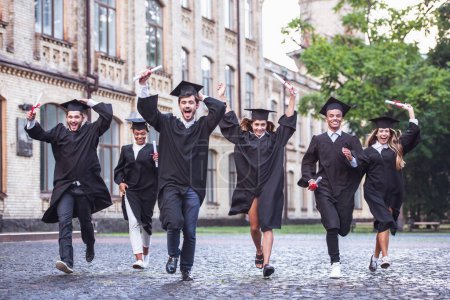 Foto de Los graduados exitosos en vestidos académicos tienen diplomas, miran a la cámara y sonríen mientras corren al aire libre - Imagen libre de derechos
