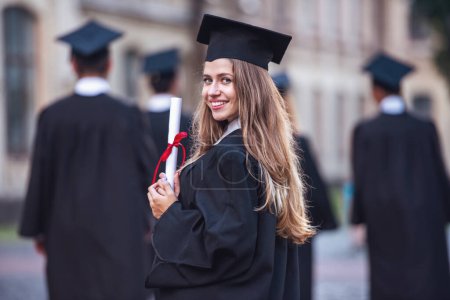 Photo for Beautiful female graduate in academic dress is holding diploma, looking at camera and smiling while standing outdoors - Royalty Free Image