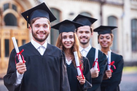 Foto de Los graduados exitosos en vestidos académicos tienen diplomas, miran a la cámara y sonríen mientras están al aire libre. - Imagen libre de derechos