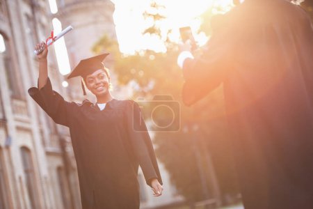 Photo for Successful graduates in academic dresses are taking photos with diplomas of each other while standing outdoors - Royalty Free Image