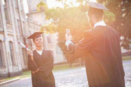 Foto de Los graduados exitosos en vestidos académicos están tomando fotos con diplomas el uno del otro mientras están al aire libre. - Imagen libre de derechos