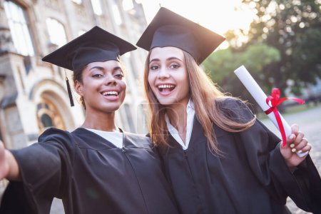 Photo for Beautiful female graduates in academic dresses are holding diplomas, looking at camera and smiling while standing outdoors - Royalty Free Image