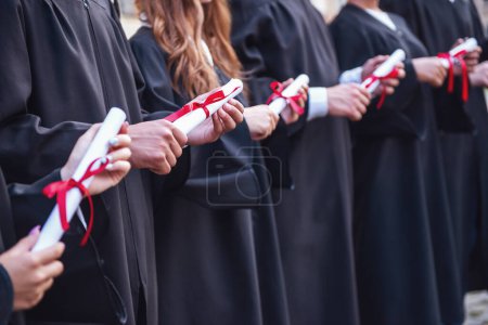Foto de Los graduados exitosos en vestidos académicos están sosteniendo diplomas, mirando hacia adelante y sonriendo mientras están de pie en una fila al aire libre - Imagen libre de derechos