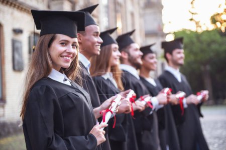 Photo for Successful graduates in academic dresses are holding diplomas, looking forward and smiling while standing in a row outdoors - Royalty Free Image