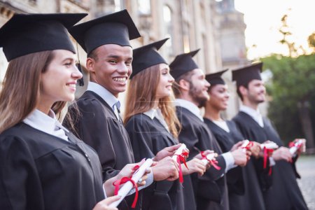Photo for Successful graduates in academic dresses are holding diplomas, looking forward and smiling while standing in a row outdoors - Royalty Free Image