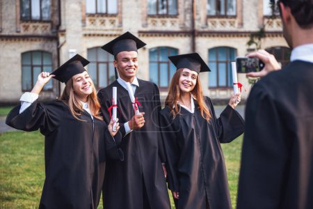 Photo for Successful graduates in academic dresses are holding diplomas, looking at camera and smiling while standing outdoors, guy is taking photo of them - Royalty Free Image