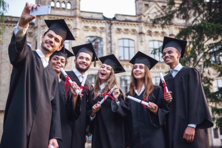 Foto de Graduados exitosos en vestidos académicos están sosteniendo diplomas, mirando a la cámara y sonriendo mientras están de pie al aire libre, chico está tomando una foto de ellos - Imagen libre de derechos