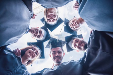 Foto de Vista inferior de graduados exitosos en vestidos académicos mirando a la cámara y sonriendo mientras están de pie al aire libre - Imagen libre de derechos