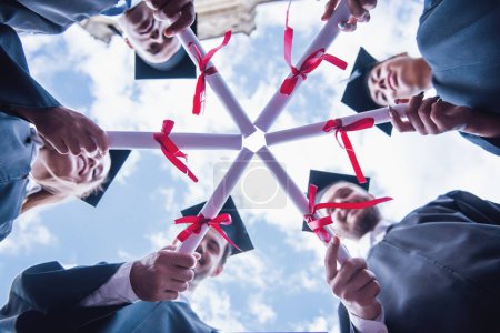 Photo for Bottom view of successful graduates in academic dresses holding diplomas and smiling while standing outdoors - Royalty Free Image