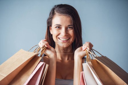 Foto de Hermosa mujer de pelo oscuro joven en vestido a rayas está sosteniendo bolsas de compras, mirando a la cámara y sonriendo, sobre fondo gris - Imagen libre de derechos