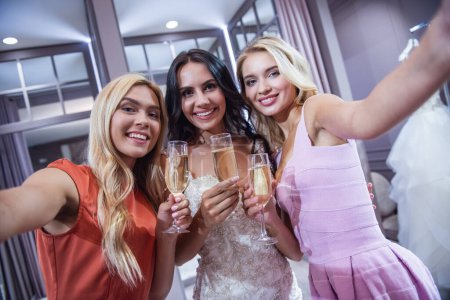 Téléchargez les photos : De belles jeunes femmes regardent la caméra, buvant du champagne et souriant tout en choisissant la robe de mariée dans le salon - en image libre de droit