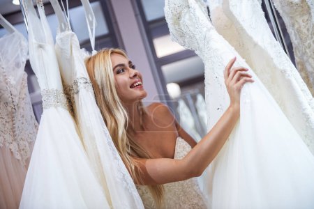 Foto de Atractiva novia joven está sonriendo al elegir el vestido de novia en el salón de bodas moderno - Imagen libre de derechos