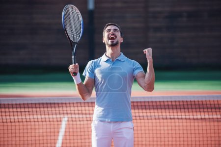 Téléchargez les photos : Beau homme tient raquette de tennis et crier de bonheur tout en jouant au tennis sur le terrain à l'extérieur - en image libre de droit
