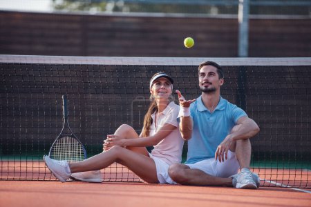 Foto de Hombre guapo y hermosa mujer están descansando en la cancha de tenis al aire libre después del juego, el hombre está lanzando una pelota - Imagen libre de derechos
