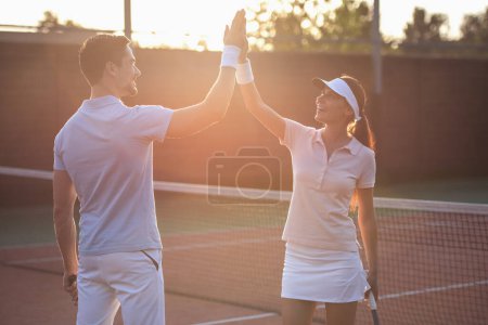 Foto de Hombre guapo y hermosa mujer están dando alta cinco y sonriendo mientras juega al tenis en la cancha de tenis al aire libre - Imagen libre de derechos
