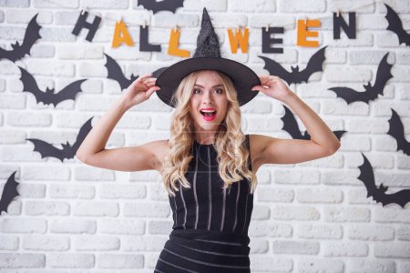 Foto de Hermosa chica rubia en vestido negro y sombrero de bruja está mirando a la cámara y sonriendo, en el fondo decorado para Halloween - Imagen libre de derechos