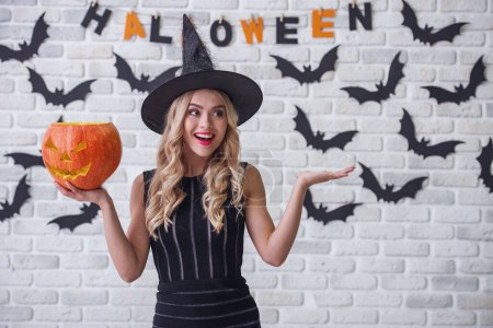 Téléchargez les photos : Belle fille blonde en robe noire et chapeau de sorcière tient une citrouille effrayante, regardant loin et souriant, sur fond décoré pour Halloween - en image libre de droit