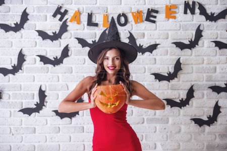 Téléchargez les photos : Belle fille sexy en robe rouge et chapeau de sorcière tient une citrouille effrayante, regardant la caméra et souriant, sur fond décoré pour Halloween - en image libre de droit