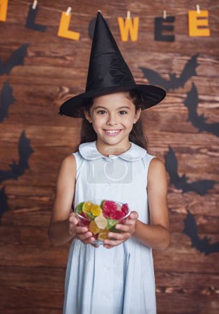 Téléchargez les photos : Petite fille mignonne en chapeau de sorcière tient un bol de bonbons, regardant la caméra et souriant, sur fond décoré pour Halloween - en image libre de droit
