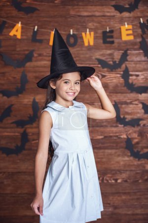 Foto de Linda niña en sombrero de bruja está mirando a la cámara y sonriendo, en el fondo decorado para Halloween - Imagen libre de derechos