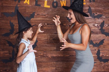 Téléchargez les photos : Petite fille mignonne et sa belle maman en chapeaux de sorcière se montrent griffes et sourire, sur fond décoré pour Halloween - en image libre de droit
