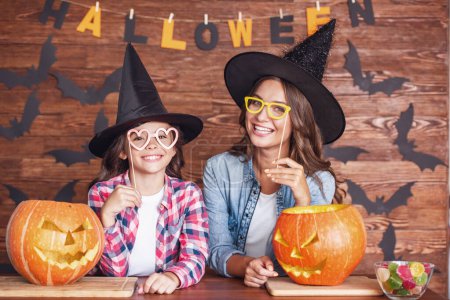 Foto de Linda niña y su hermosa mamá en sombreros de bruja están mirando a la cámara y sonriendo mientras se sienta en el fondo de madera decorado para Halloween - Imagen libre de derechos