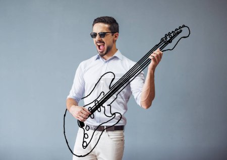 Téléchargez les photos : Beau jeune homme en vêtements décontractés intelligents et lunettes de soleil imite jouer de la guitare et chanter, sur fond gris - en image libre de droit