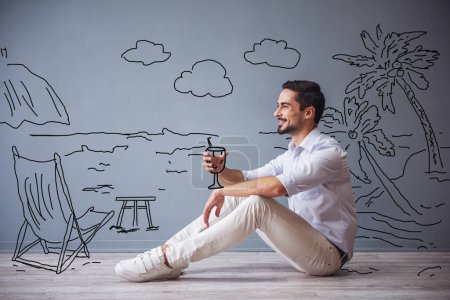 Téléchargez les photos : Beau homme en vêtements décontractés intelligents imite tenant un cocktail et souriant tout en étant assis sur le sol à côté du mur avec dessiné bord de mer - en image libre de droit