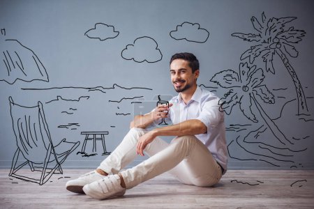 Téléchargez les photos : Beau homme en vêtements décontractés intelligents imite tenant un cocktail et souriant tout en étant assis sur le sol à côté du mur avec dessiné bord de mer - en image libre de droit