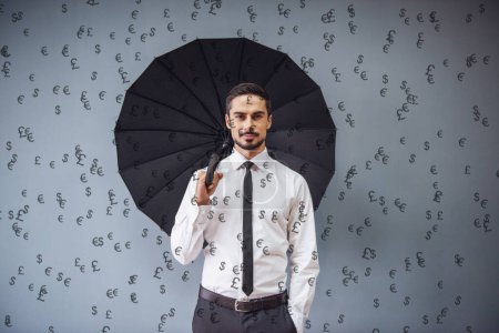 Foto de Hombre de negocios guapo en traje clásico con paraguas está mirando a la cámara, los billetes sorteados están cayendo como la lluvia, sobre un fondo gris - Imagen libre de derechos