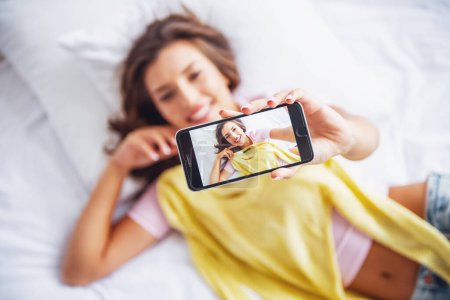 Téléchargez les photos : Belle fille en vêtements décontractés fait selfie avec un chemisier en utilisant un téléphone intelligent et souriant tout en étant couché sur le lit à la maison - en image libre de droit