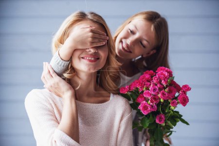 Foto de Bonita hija adolescente sostiene flores y cubre los ojos de su madre mientras hace una sorpresa, ambos sonríen - Imagen libre de derechos