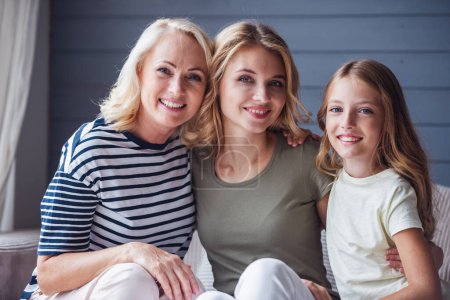 Photo for Beautiful women generation: granny, mom and daughter are hugging, looking at camera and smiling while sitting on couch at home - Royalty Free Image