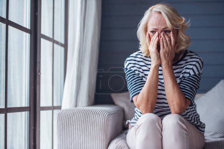 Foto de Hermosa mujer mayor triste está llorando y limpiando sus lágrimas mientras está sentada en el sofá en casa - Imagen libre de derechos