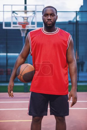 Téléchargez les photos : Beau joueur de basket est debout avec une balle sur le terrain de basket à l'extérieur - en image libre de droit