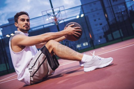 Photo for Handsome basketball player is sitting with a ball on basketball court outdoor in the evening - Royalty Free Image