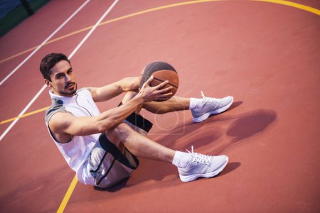 Photo for Handsome basketball player is looking at camera while sitting with a ball on basketball court outdoors - Royalty Free Image