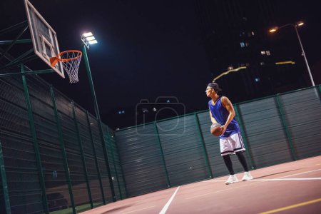 Téléchargez les photos : Portrait complet d'un jeune joueur de basket élégant en casquette sautant et tirant une balle à travers le cerceau tout en jouant à l'extérieur la nuit - en image libre de droit