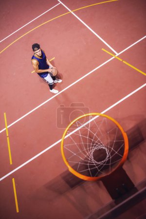 Téléchargez les photos : Vue du dessus du jeune joueur de basket élégant en casquette tirant une balle à travers le cerceau tout en jouant à l'extérieur la nuit - en image libre de droit