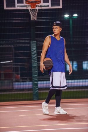Téléchargez les photos : Portrait complet de jeune joueur de basket élégant en casquette debout avec une balle sur le terrain de basket-ball en plein air la nuit - en image libre de droit