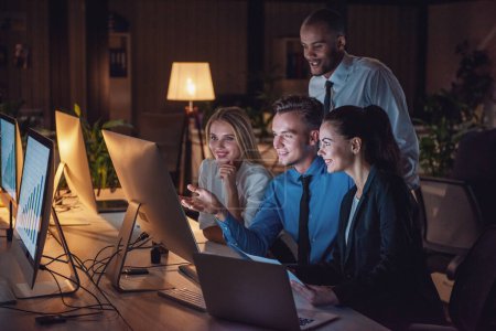 Foto de La gente de negocios exitosa está usando una computadora y sonriendo mientras trabaja en la oficina hasta altas horas de la noche - Imagen libre de derechos