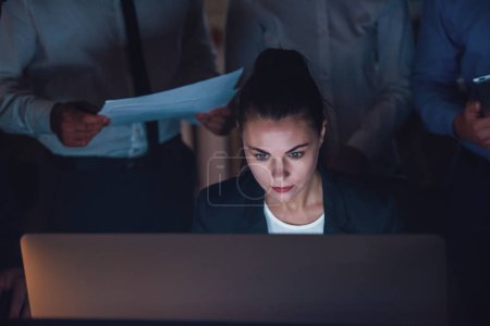 Téléchargez les photos : Belle femme d'affaires utilise un ordinateur tout en travaillant au bureau tard dans la nuit, collègues sont debout autour - en image libre de droit