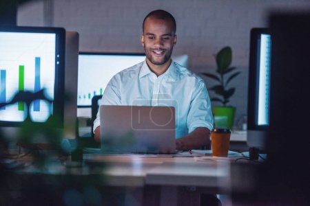 Foto de Hombre de negocios afroamericano guapo en traje clásico está usando un ordenador portátil y sonriendo mientras trabaja en la oficina hasta tarde en la noche - Imagen libre de derechos