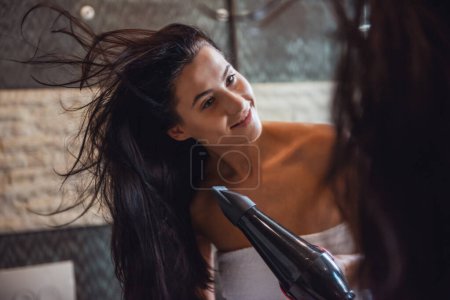 Photo for Attractive young woman in towel is drying her hair and smiling while looking into the mirror in bathroom - Royalty Free Image