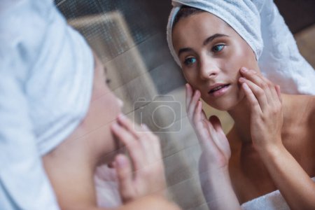 Foto de Mujer joven con toalla en la cabeza está tocando sus mejillas mientras se mira al espejo en el baño - Imagen libre de derechos