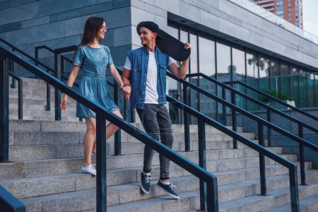 Photo for Stylish young couple is holding hands and smiling while walking outdoors, guy is holding a skateboard - Royalty Free Image