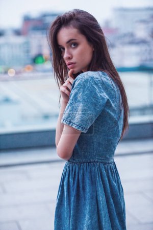 Téléchargez les photos : Portrait de belle fille brune en robe de jean regardant la caméra, sur le fond de la ville - en image libre de droit